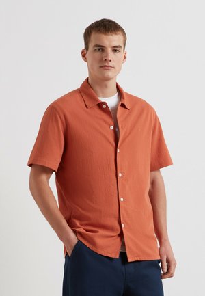 Młody mężczyzna stoi z rękami w kieszeniach, ubrany w pomarańczową koszulę z krótkim rękawem na guziki, na białej koszulce, oraz w granatowe spodnie.