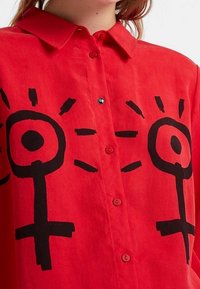 Czerwona bawełniana koszula z dużymi czarnymi feministycznymi symbolami, zapinana na guziki z kołnierzykiem w szpic oraz krótkimi rękawami o gładkiej fakturze.