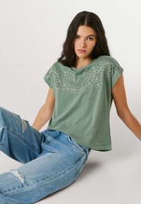 Νεαρή γυναίκα με μακριά σκούρα μαλλιά, ντυμένη με πράσινη κεντημένη μπλούζα με κοντά μανίκια και σκισμένο μπλε τζιν, καθισμένη μπροστά σε απλό φόντο.