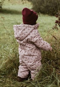 Roze gewatteerde sneeuwpak met kleine kerspatronen, elastische tailleband en een capuchon. Gecombineerd met een bordeauxrode gebreide muts. Grasachtergrond.