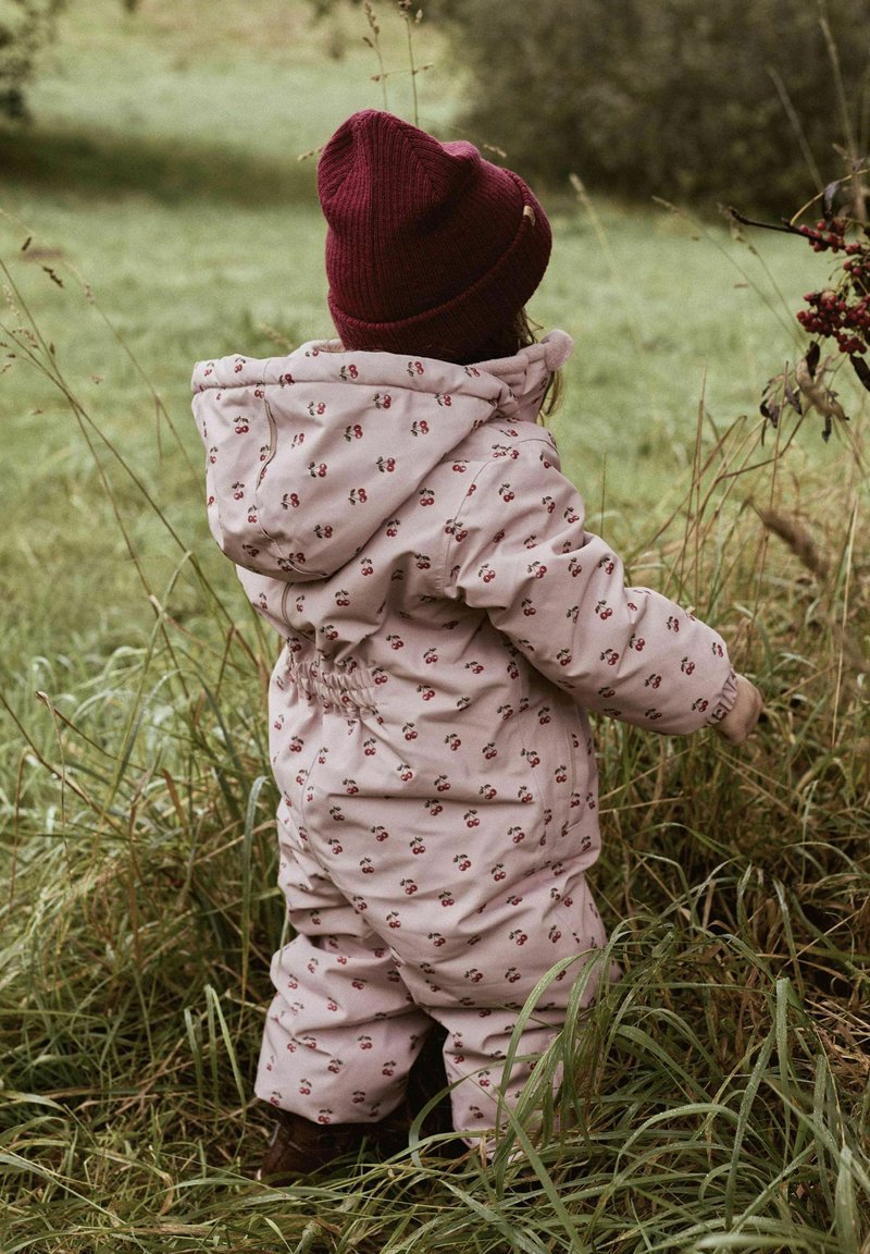 Roze gewatteerde sneeuwpak met kleine kerspatronen, elastische tailleband en een capuchon. Gecombineerd met een bordeauxrode gebreide muts. Grasachtergrond.