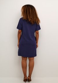 Kortärmad klänning i mörk marinblå, knälång, tillverkad av textil med struktur, rak snitt och sidofickor. Bruna klackar bärs.