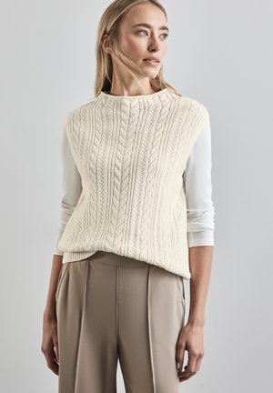 Strukturierte cremefarbene Kabelstrickweste mit hohem Kragen, über einem weißen Langarmshirt getragen, kombiniert mit beigen hoch-taillierten Hosen.