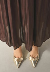 Metāliskas zelta uzpirksteņu kurpes ar loku akcentu, apvienotas ar plūstošu melnu puscaurspīdīgu svārku, kas pieskaras gludai pelēkai virsmai.