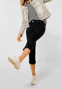 Pantaloni neri a vita alta, taglio slim, abbinati a una blusa con motivo bianco e nero e un giubbotto grigio chiaro. Le calzature sono sneakers bianche.