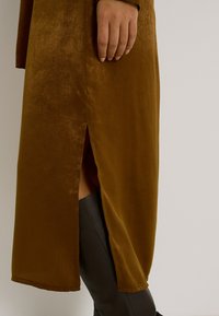 Vestido de terciopelo marrón con una abertura lateral, que presenta una textura suave y un corte relajado. Botas negras hasta la rodilla visibles por debajo.