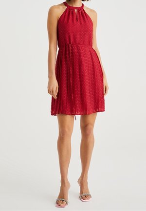 Mujer con vestido sin mangas rojo texturizado de tipo halter con lazo en la cintura y sandalias de punta abierta, de pie sobre fondo blanco.