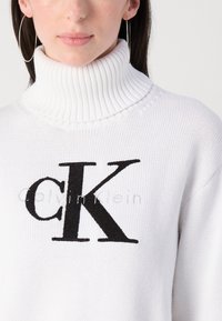 Hvit strikket højhalset genser med et fremtrædende sort "CK" logo. Tekstureret stof med ribbet kravedetaljer. Subtil branding under logoet.