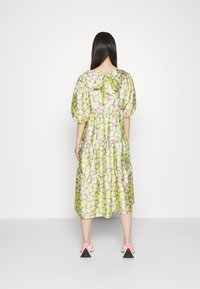Femme aux longs cheveux foncés portant une robe midi fleurie vert clair et violet avec des manches bouffantes et un nœud noué dans le dos, associée à des chaussures roses et blanches.