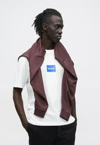 Man met dreadlocks draagt een wit Hugo T-shirt, een bordeauxrode trui over zijn schouders en zwarte broek, kijkt naar rechts.