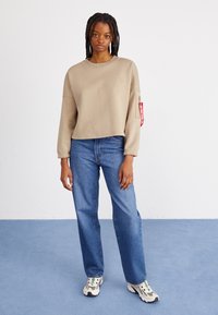 Beige cropped sweatshirt with long sleeves, elastic cuffs, and a side pocket. Paired with blue straight-leg jeans and multi-colored sneakers.