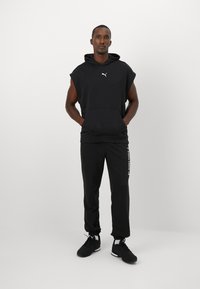 Zwart mouwloos hoodie met kangoeroezak en logo, gecombineerd met zwarte taps toelopende broek met witte opdruk, gedragen met zwarte sportschoenen.