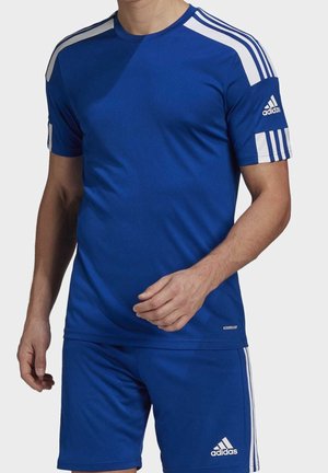 Homme portant une tenue de sport Adidas bleue avec des rayures blanches sur les épaules et le short, debout devant un fond clair uni.