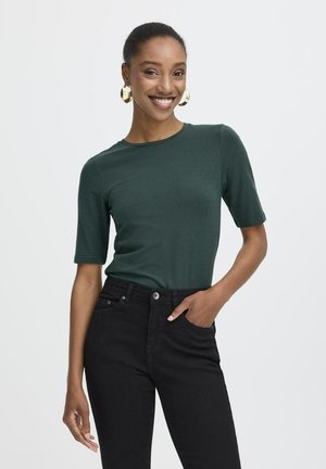 Femme souriante avec des boucles d'oreilles créoles dorées porte un haut à manches courtes vert foncé et un jean noir taille haute, main dans la poche, sur un fond uni.