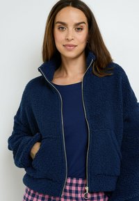 Donna che indossa una giacca in pile blu navy con zip, un top blu navy e pantaloni a scacchi rosa e blu navy, in piedi davanti a uno sfondo bianco semplice.