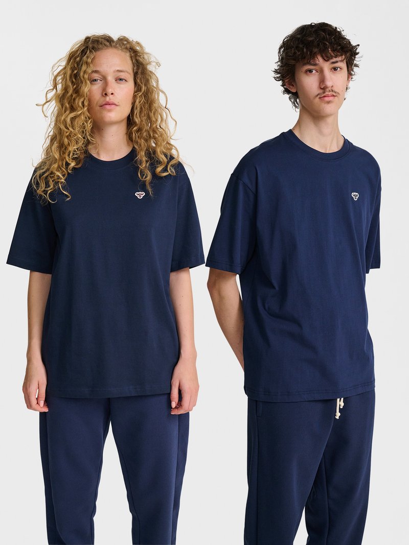 Zwei junge Erwachsene, die passende dunkelblaue T-Shirts und Hosen tragen, stehen vor einem einfachen weißen Hintergrund und schauen nach vorne.