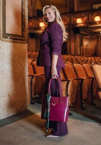 Vínová taška přes rameno s texturovaným krokodýlím akcentem, držená s šátkem se zeleným vzorem, doplněná ladícím fialovým oblekem v divadelním prostředí.
