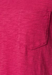 Koszula z krótkim rękawem w jasnoróżowym kolorze, wykonana z teksturowanej tkaniny. Posiada małą kieszeń na piersi po lewej stronie z detalami w postaci przeszyć.