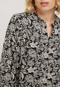 Blouse à imprimé floral avec un fond noir et des motifs blancs complexes, un col montant et une patte de boutonnage. Coupe décontractée avec des manches longues.