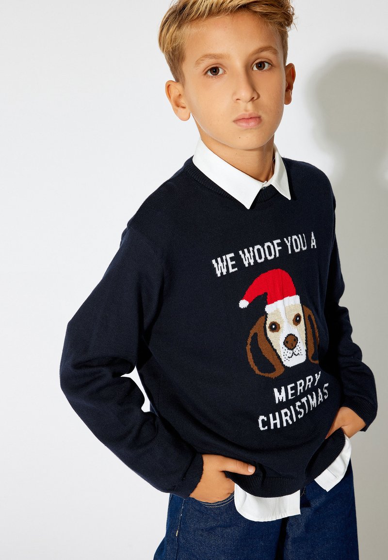 Granatowy sweter dziany z grafiką beagle'a w czerwonej czapce świętego Mikołaja oraz napisem "ŻYCZYMY CI WESOŁYCH ŚWIĄT". Biała koszula z kołnierzykiem pod spodem.