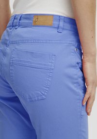 Modré bavlněné kalhoty s hladkou texturou, se zadní kapsou a hnědou koženou štítkem na pase. Boční kapsa je viditelná.