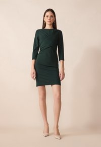 Temnozelena ovita obleka s tridelnimi rokavi, prilegajoč design in razporkoma na robu, v kombinaciji s svetlimi čevlji s koničastim prstom.