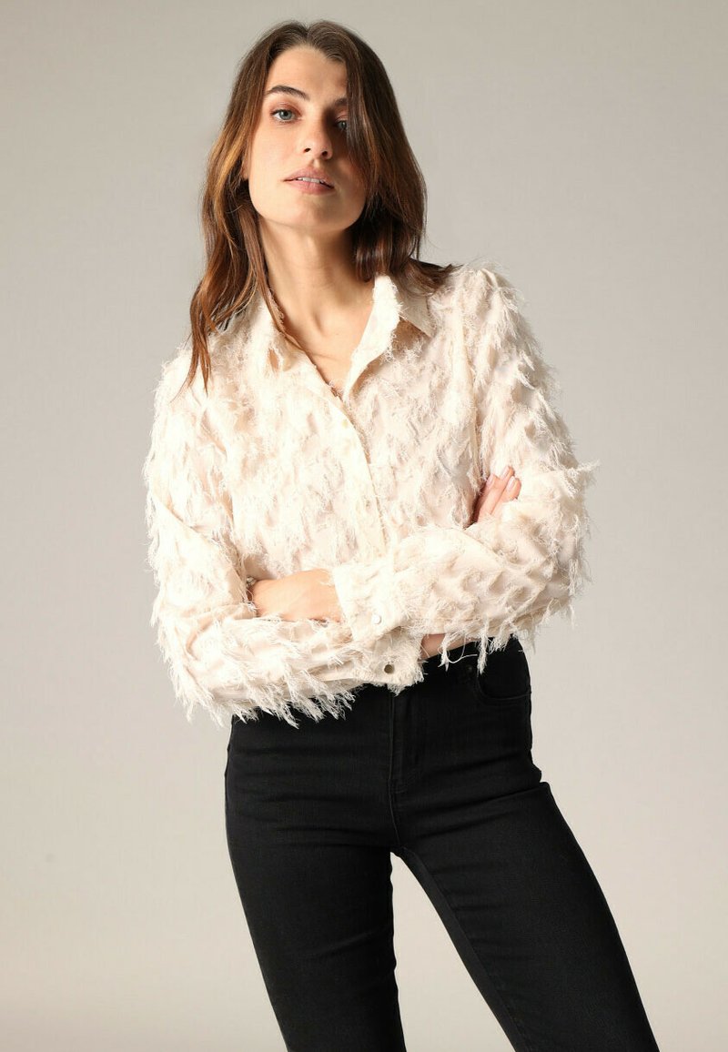 Vrouw met lang bruin haar die een gemêleerd crèmekleurig blouse met knopen en zwarte high-waisted broek draagt, met gekruiste armen.