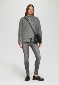 Veste en mélange de laine grise avec un devant à boutons, superposée sur un col roulé gris clair. Associée à un jean gris foncé et des mocassins noirs.