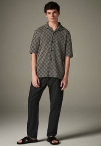 Camicia a maniche corte con motivi bianchi e neri e colletto, abbinata a pantaloni neri e sandali neri, in piedi contro uno sfondo neutro.