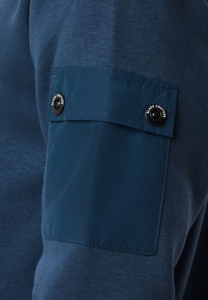 Granatowy sweter z teksturowanej tkaniny, z kontrastową gładką łatą i detalem napy na lewym rękawie.