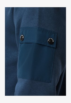 Granatowy sweter z teksturowanej tkaniny, z kontrastową gładką łatą i detalem napy na lewym rękawie.