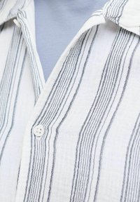 Biela košeľa na gombíky s tenkými čiernymi vertikálnymi pruhmi, nosená cez svetlomodré tričko.