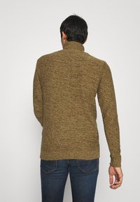 Homme portant un pull à col roulé marron tricoté et un jean bleu foncé, debout de dos face au spectateur sur un fond uni.