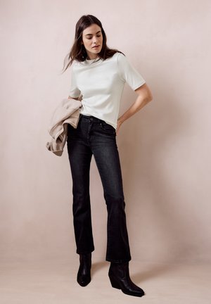 Femme portant une chemise blanche à manches courtes, un jean noir taille haute, des bottines noires, tenant une veste beige sur un fond beige uni.