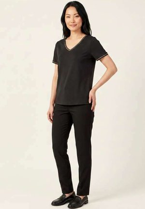 Femme aux longs cheveux noirs portant un haut noir à col en V manches courtes, un pantalon noir et des chaussures noires sans lacets, debout avec une main sur la hanche.