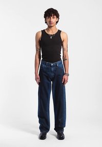 Svart linne, vida mörkblå jeans med silverdetaljer på byxorna, bärs med svarta skor. Modellen står upprätt mot en vit bakgrund.