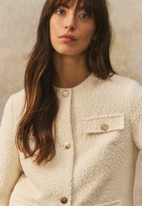 Texturierte Cremejacke mit goldenen, strukturierten Knöpfen, Rundhalsausschnitt und einer kleinen Brusttasche. Geeignet für verschiedene Anlässe.