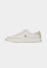 Sneaker bas blanc et beige avec lacets à l'avant et logo "cK" sur le côté, présenté sur un fond clair uni.