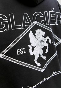 Sudadera negra con un gráfico blanco de un caballo alado dentro de una forma de diamante, con el texto "GLACIÉR" y "EST. 19" en letras blancas negritas.