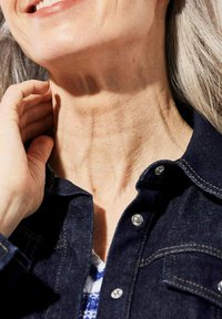 Donkerblauwe denim jas met zilveren drukknoopjes, met een gestructureerde neklijn en grijs haar in fel licht. Huid vertoont zichtbare textuur.