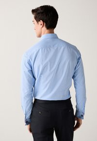 Šviesiai mėlyna ilgomis rankovėmis marškiniai su tekstūruotu raštu, kontrastingais mygtukais ant rankogalių ir klasikiniu apykakliu. Dėvimi su tamsiomis kelnėmis.