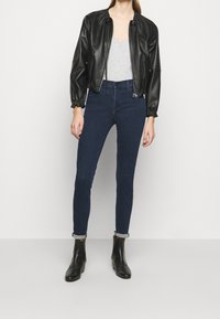 Chaqueta bomber de cuero negro con puños fruncidos, camiseta gris con escote en V, jeans skinny oscuros y botas negras de tobillo con tacones apilados.