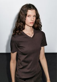 T-shirt marrone a scollo a V, maniche corte, design aderente, texture liscia. Il modello indossa una collana in argento. Lo sfondo presenta superfici bianche e scure.