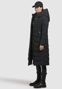 Lungo cappotto imbottito nero con colletto alto e tasche con cerniera laterali. Presenta un motivo a zig-zag trapuntato e un tirante della cerniera con marchio. Indossato con stivali neri alla caviglia.