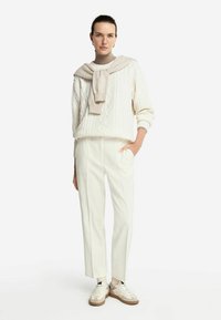 Weißer Kabelstrickpullover, hellbeige Schal über den Schultern drapiert, maßgeschneiderte weiße Hose und weiße Sneakers mit sandfarbenen Akzenten.