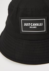 Cappello a secchiello nero fatto di tessuto con un'etichetta di marca ben visibile. Presenta una visiera larga e flessibile e cuciture testurizzate lungo i lati.