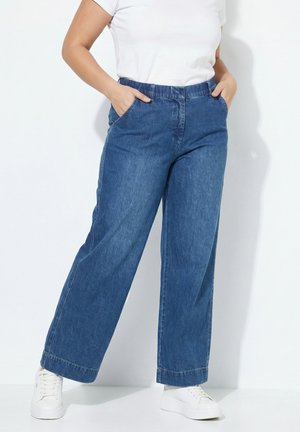 Person trägt blaue Jeans mit weitem Bein, weiße Sneakers und ein weißes kurzärmliges Hemd, steht mit Händen in den Taschen vor einem weißen Hintergrund.