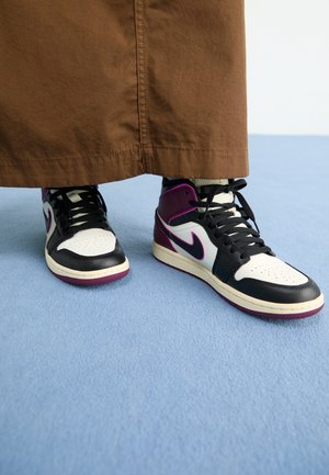 Osoba nosząca brązowe spodnie i czarne, białe oraz fioletowe wysokie sneakersy Nike stoi na jasnoniebieskiej podłodze.