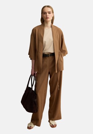 Vrouw die bruine wijde broek draagt, bijpassend open jasje, beige top, een donkerbruine tas vasthoudend en crèmewitte geweven sandalen, staand tegen een witte achtergrond.