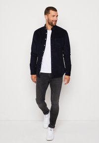 Marinblå kordflorsskjorta med knapp, utrustad med en bröstficka. Skjortan är över en vit T-shirt och kombineras med mörka jeans och vita sneakers.